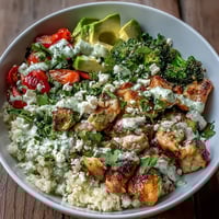 Golden seared chicken sits atop fluffy cauliflower rice with bright red bell peppers and avocado slices.