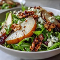 Fresh arugula and pear salad with creamy cheese, crunchy nuts, and tangy balsamic dressing in a white bowl.